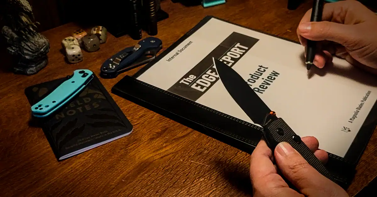 A person is holding a Vosteed Raccoon in their left hand, a Uni-ball zento signature pen in the right hand. There are knives and flashlights laying on the desk. A report lays on the desk that is titled The Edge Report with text below that says Product Review. The top of the report says Internal Document and the bottom says A Magnolia Blade Publication