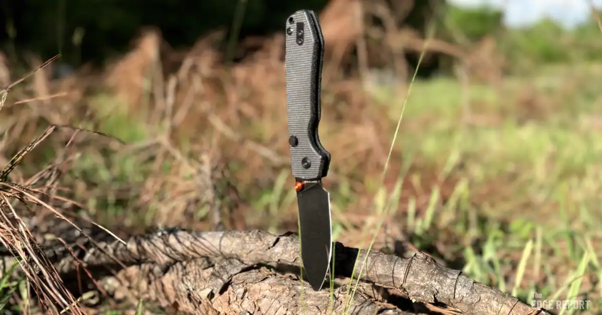 A Vosteed Raccoon with a black stonewashed blade, brown micarta scales and an orange thumb stud stuck in a brand with a blurred field in the background.
