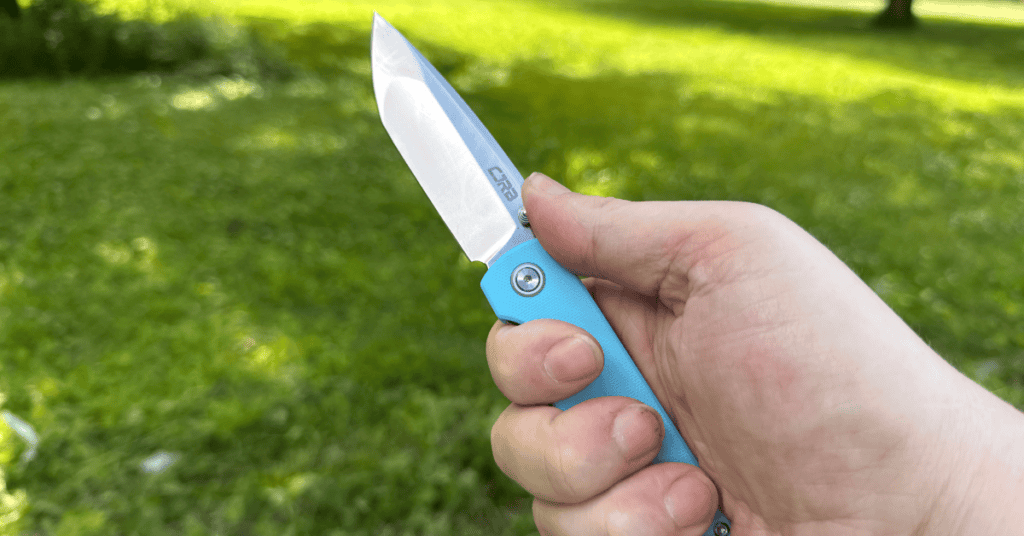 Full grip on the CJRB Nova folding knife with baby blue G10 handle, open blade, green backyard in the background